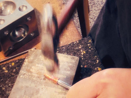 Gros plan sur une main frappant une tige de cuivre avec un marteau, posée sur une plaque d’acier. La scène, prise en atelier, capture le geste en mouvement, entre force et précision. En arrière-plan, une matrice métallique et les textures du plan de travail suggèrent l’univers artisanal, brut et vivant.
