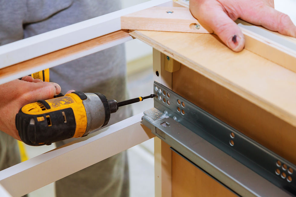 Person using a yellow drill to assemble white wooden cabinets with drawer glides. Close-up, indoor setting. Hands focused on work.