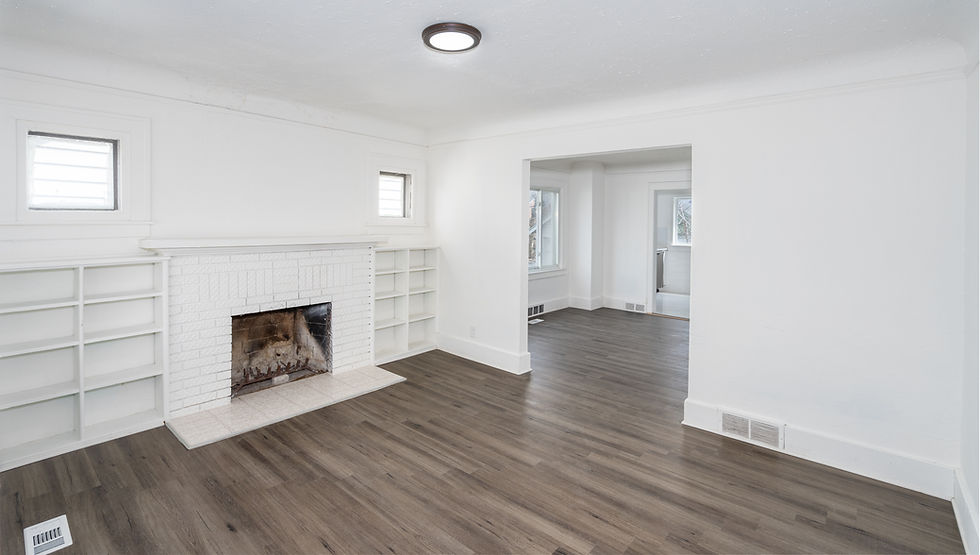Empty room with white walls, a white brick fireplace, built-in shelves, and dark wooden floors. Natural light from windows, modern light fixture.