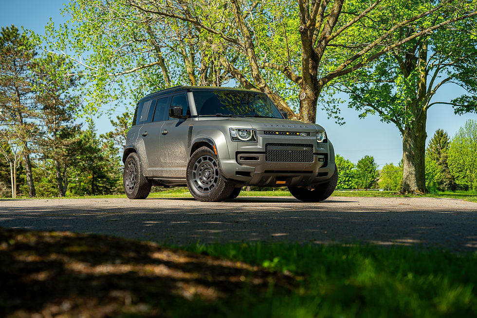 Defender OCTA parked on a sunny road with lush green trees in the background. Bright daylight highlights its sleek design and rugged tires.
