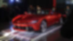 2026 Ferrari Amalfi Spider - Red Ferrari convertible on display with people observing, dimly lit event setting, spotlighting on car, text reads Ferrari.