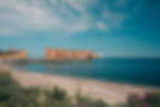 Coastal view with a red-roofed house on a cliff, rock formations in blue sea, pebble beach, and flowers under a partly cloudy sky. Serene mood. Percé Rock