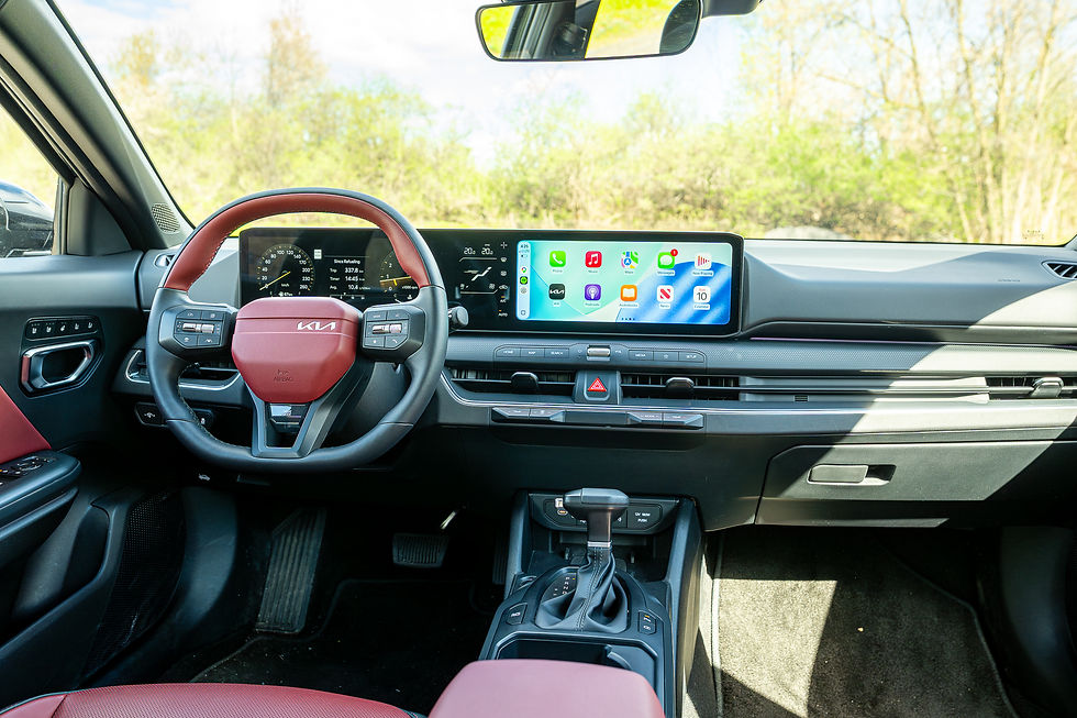 2026 KIA K4 - Kia car interior with red steering wheel and dual digital displays, parked in bright daylight with trees outside.