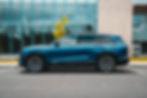 Cadillac Vistiq is parked in front of a glass building with trees reflecting. Clear skies and a yellow-leaved tree nearby. Building number 22 visible.