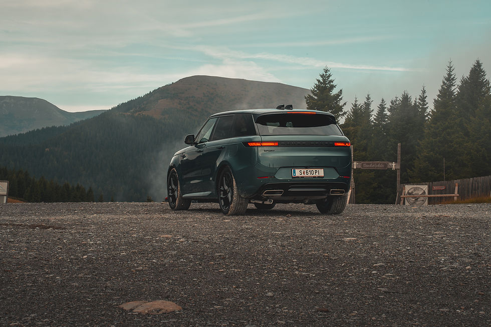 Range Rover Sport P460e dark green on a gravel road with forested mountains in the background. Overcast sky, peaceful mood.