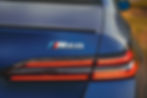 2026 BMW i5 M60 - Close-up of a blue car's rear, featuring "M60" badge. Red tail lights illuminate. Background shows a blurred outdoor setting.