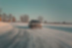 2026 Volvo EX 30 Cross Country A black car drifts on snowy terrain with blurred snow trails. Trees line the horizon under a blue sky. Headlights are on.