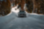 SUV driving on a snowy road through a forest. Snowbanks line the path, and tall trees create a backdrop. The scene is serene and wintry.
