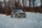 2026 Volvo XC90 T8 - Silver SUV driving through a snowy forest road, surrounded by bare trees. The scene is calm with a clear blue sky overhead.