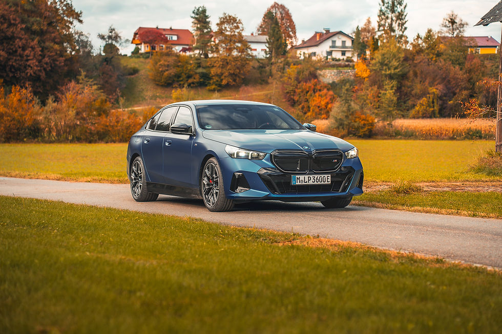 2026 BMW i5 M60 - Blue car on a rural road, surrounded by autumn trees and distant houses. Sunny day with vibrant colors; license plate reads "M LP3600E".