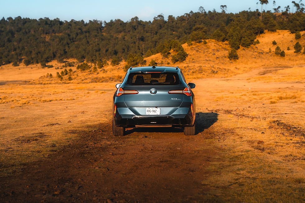 2026 BMW X3 xDrive30e - A blue SUV drives on a dirt path in a golden, grassy landscape with a forested hill in the background. Sunlight casts warm tones.