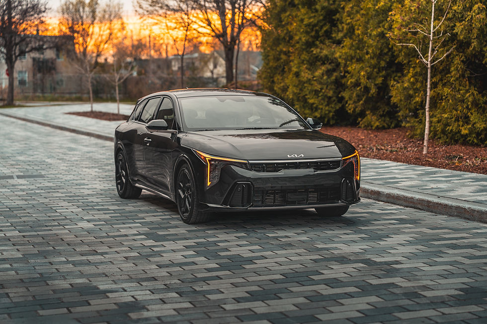 2026 KIA K4 - Black Kia sedan parked on a brick road at sunset, with amber headlights on and trees and homes in the background.