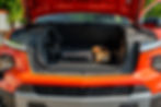 Chevrolet Silverado EV Open frunk of an orange vehicle with a black bag and a cardboard box inside. Green foliage in the background, sunlight illuminating the scene.