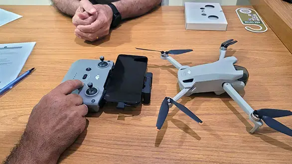 Um controle remoto de drone com um smartphone acoplado e um drone cinza com hélices abertas estão sobre uma mesa de madeira, junto a documentos, indicando um ambiente de estudo e prática.