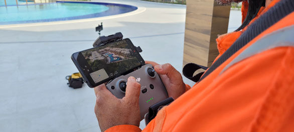 Mãos de um operador da Drone Santa Catarina, vestindo uniforme laranja, controlando um drone com um smartphone acoplado, que exibe uma vista aérea de um complexo de edifícios e piscinas em Santa Catarina. Uma piscina azul está em primeiro plano.