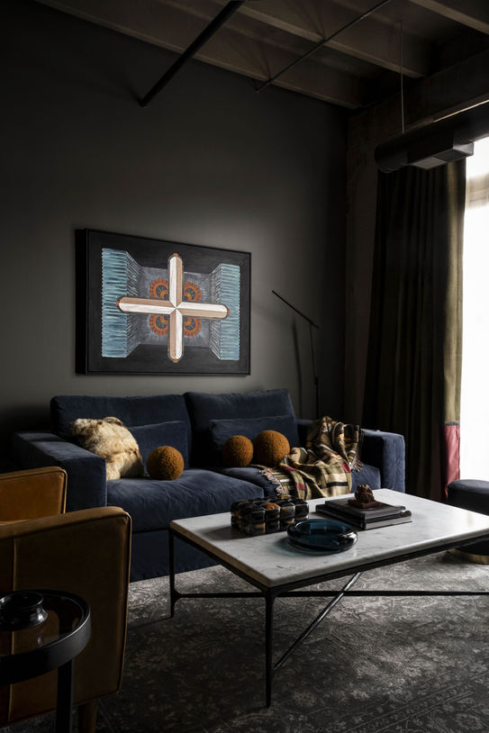 A moody living room with a deep blue velvet sofa, rust-colored accent pillows, and a modern abstract artwork above. The space features layered textures, a marble coffee table with decor, and dark walls that create a dramatic loft aesthetic.