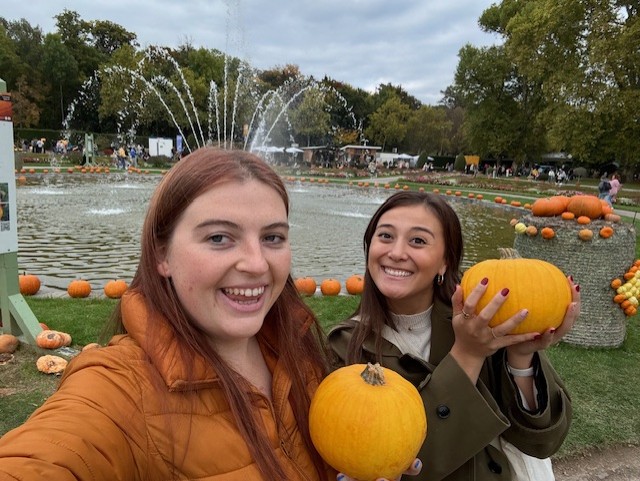 2 girls smiling in front of a fountain at the pumpkin patch holding two small pumpkins