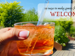 A hand holding a bright orange summer drink outdoors on a sunny patio with greenery in the background
