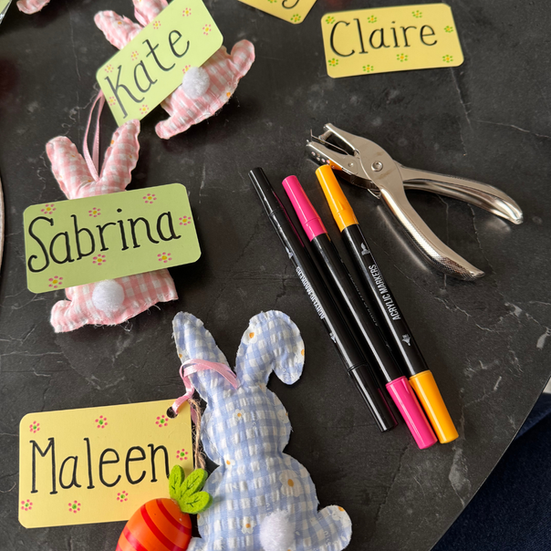 Personalized Easter place cards, bunny decorations, colorful markers, and a hole punch arranged for spring table setting preparation.