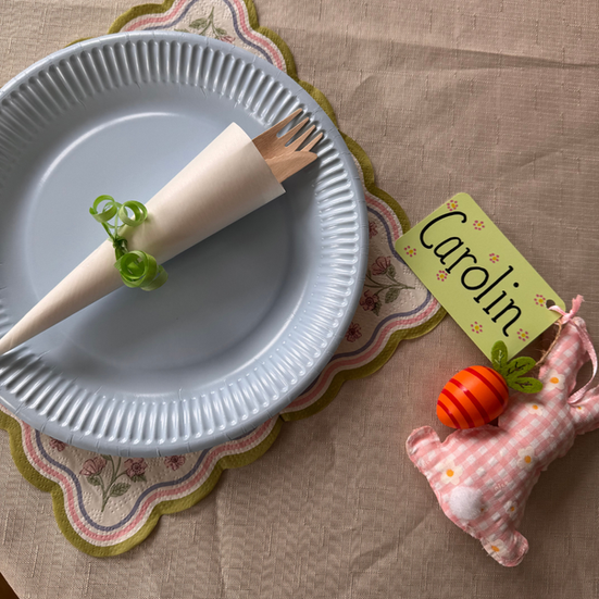 Easter-themed table setting featuring a blue plate, folded napkin with wooden cutlery, floral scalloped placemat, and a personalized bunny-shaped place card.