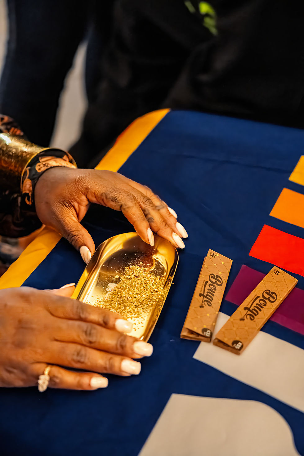 A close-up high-angle shot of a person's hands preparing to roll cannabis. The hands are holding a small gold tray filled with ground green herb. Two packages of Bouqé brand rolling papers sit on the deep blue tablecloth next to the tray.