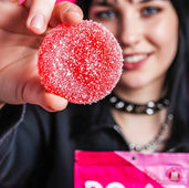 A vertical lifestyle photograph featuring a woman with dark hair and a black choker against a vibrant pink background. She is holding a single, sugar-coated red watermelon gummy in sharp focus in the foreground, while her other hand holds a pink Do Drops Focus 1:1:1 CBG:THC:THCV package. The lighting is high-energy and bright, with the woman's facial features softly blurred in the background to maintain focus on the product texture.