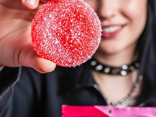 A vertical lifestyle photograph featuring a woman with dark hair and a black choker against a vibrant pink background. She is holding a single, sugar-coated red watermelon gummy in sharp focus in the foreground, while her other hand holds a pink Do Drops Focus 1:1:1 CBG:THC:THCV package. The lighting is high-energy and bright, with the woman's facial features softly blurred in the background to maintain focus on the product texture.