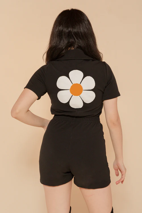 Woman wearing a black romper with a large white daisy graphic on the back, featuring an orange center.
