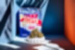 A photograph of Cake Butta cannabis packaging with vibrant colors and bold branding, accompanied by a large cannabis bud resting on a sculpted white pedestal. The scene is lit with dramatic shadows and features a mix of blue, orange, and neutral tones for a contemporary, artistic feel.