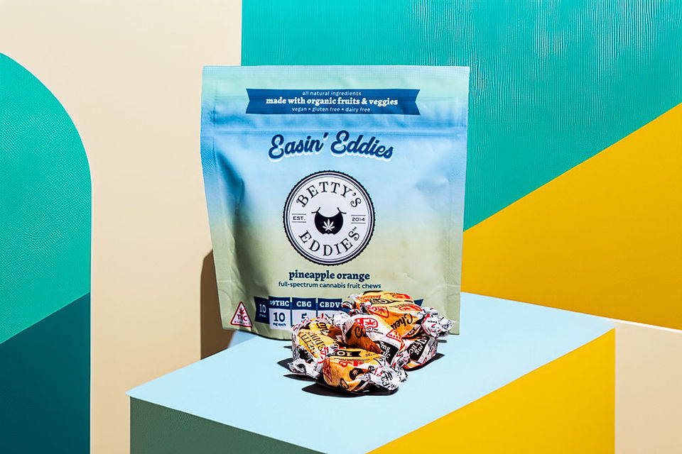 Betty's Eddies Easin' Eddies Pineapple Orange taffy bag on a colorful geometric background, with individually wrapped candies in the foreground. 