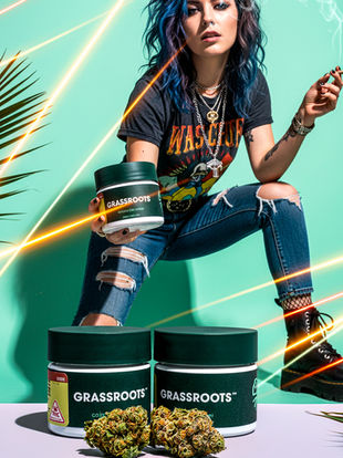 A woman with blue hair and ripped jeans crouches in the background. In the foreground, two Grassroots cannabis jars and detailed cannabis flower buds sit on a surface with laser effects.