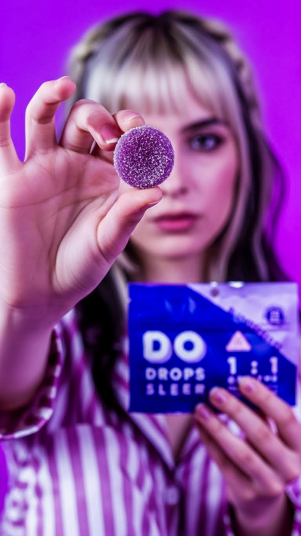 A vertical, cinematic lifestyle photograph featuring a woman with blonde hair in pink and white striped pajamas against a vivid, solid purple background. She holds a single, purple sugar-coated gummy between her thumb and forefinger in a sharp focus in the foreground. In her other hand, she holds a blue and purple Do Drops Sleep 1:1 CBN:THC package.