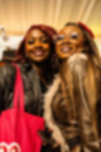 Close-up portrait of two women smiling at the event. The woman on the left wears a leather jacket, and the woman on the right wears stylish glasses and a fur-trimmed jacket.