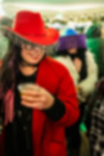 A guest wearing a red cowboy hat with rhinestone fringe holding a drink, standing in front of another guest wearing a purple fringe hat.