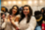 Two women in white outfits posing for a photo; the woman on the left wears a white sequin dress and holds a pre-roll, while the woman on the right wears a pearl-studded sweater.