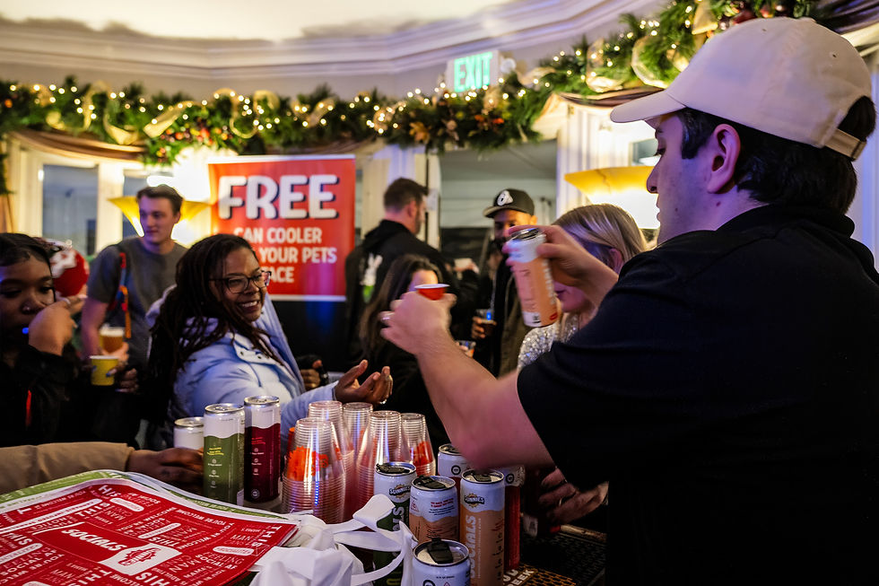 A close-up of a pouring station where a man in a white cap pours a canned beverage into small tasting cups. A red sign in the background reads "FREE Can Cooler".