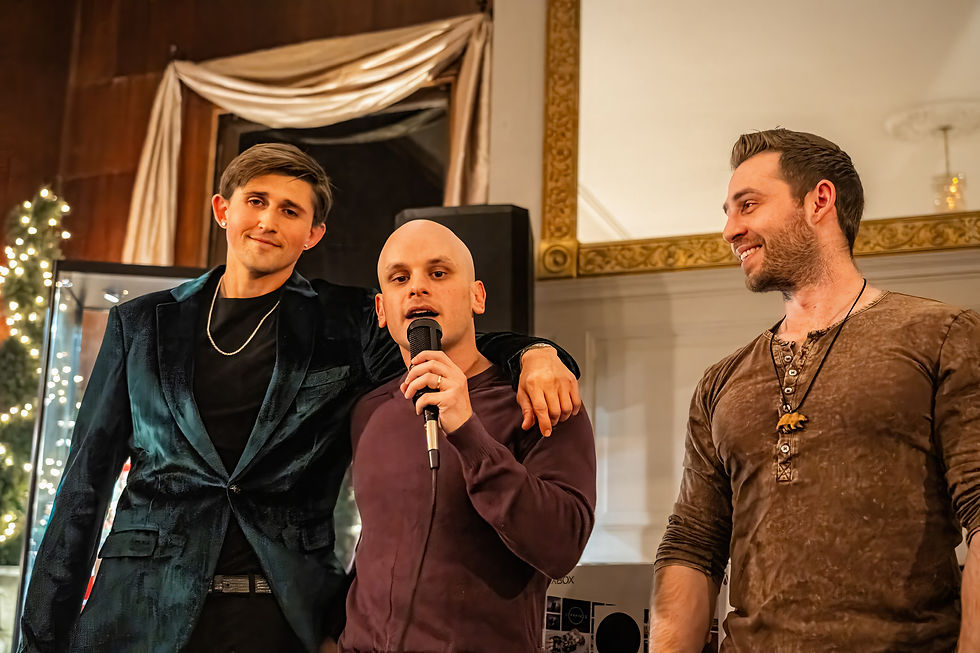 Three men standing together on stage. The man in the center is speaking into a microphone while the man in the teal velvet blazer has his arm around him. The mood is supportive and attentive.