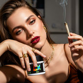 A woman with bold makeup and gold jewelry poses with Curio Wellness OG Kush Breath, holding a lit joint above a marble table covered with premium cannabis buds.