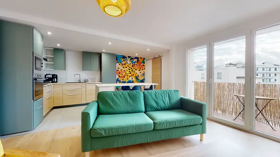 Salon avec un canapé vert émeraude, une cuisine ouverte aux tons bleu-gris et  clair, , de grandes fenêtres donnant sur un balcon avec une balustrade en bambou, et un parquet clair.
Colocation Jungle à Annemasse