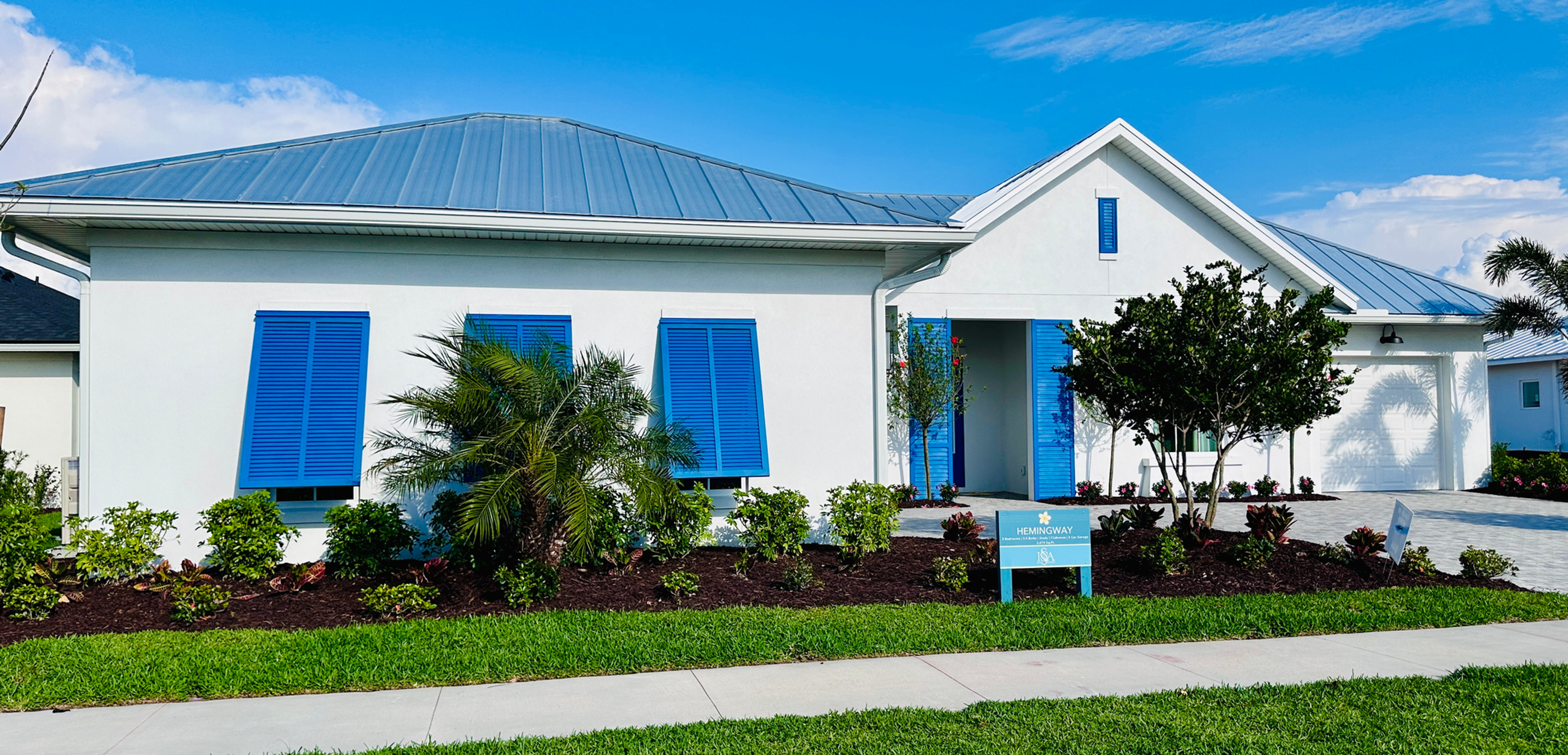 White house with blue shutters and roof, surrounded by green shrubs and a palm tree. A sign in front reads "Hemingway." Clear blue sky.