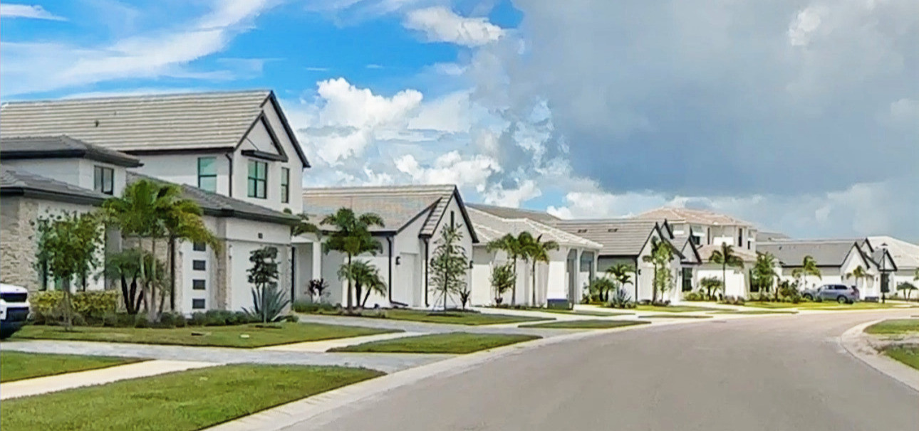Curving suburban street with white modern houses, palm trees, and a bright blue sky with large clouds.