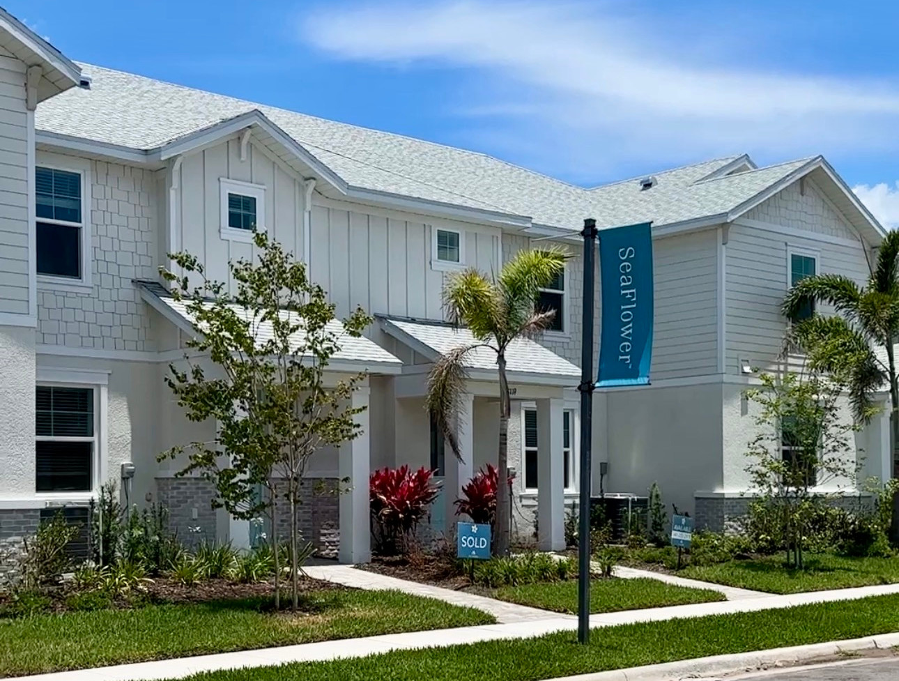 Sunny suburban townhouse with a blue Seaflower banner and a SOLD sign in the front yard, framed by palm trees.