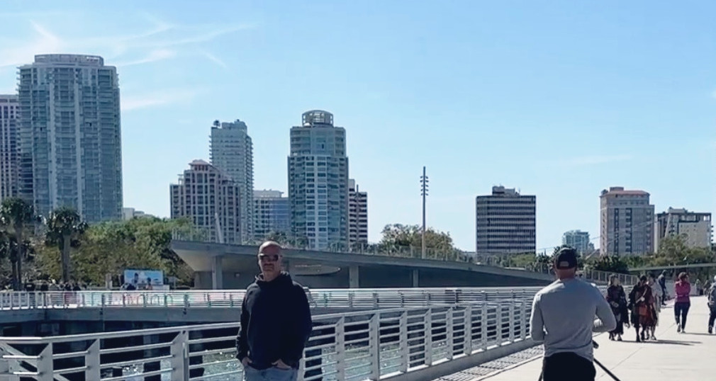 People walking on a sunny boardwalk with dogs, surrounded by palm trees and high-rise buildings. A relaxed, urban atmosphere.