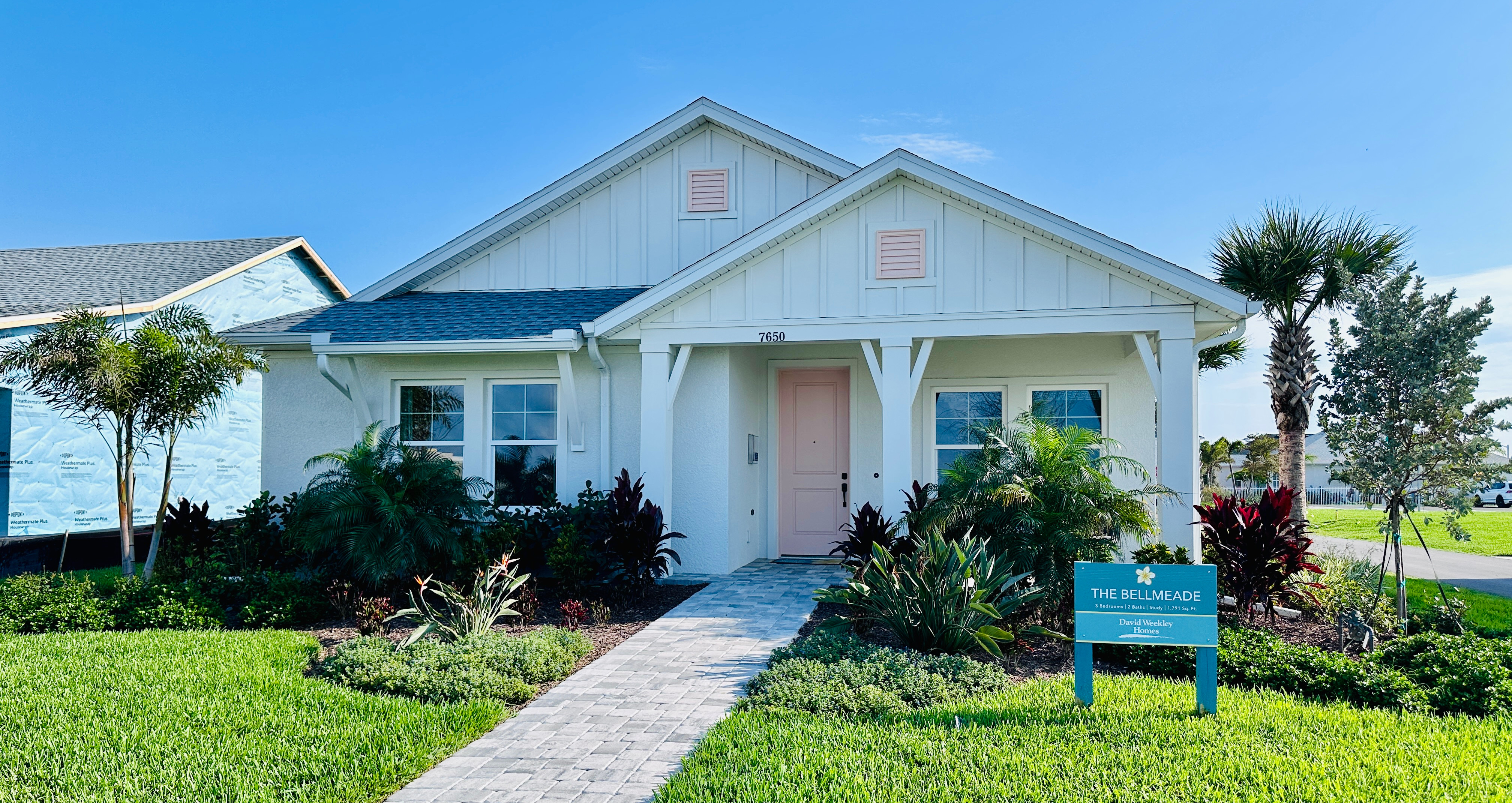 White house with gray roof, pink door, surrounded by green lawn and palm trees. Sign reads "The Bellmeade." Clear blue sky above.