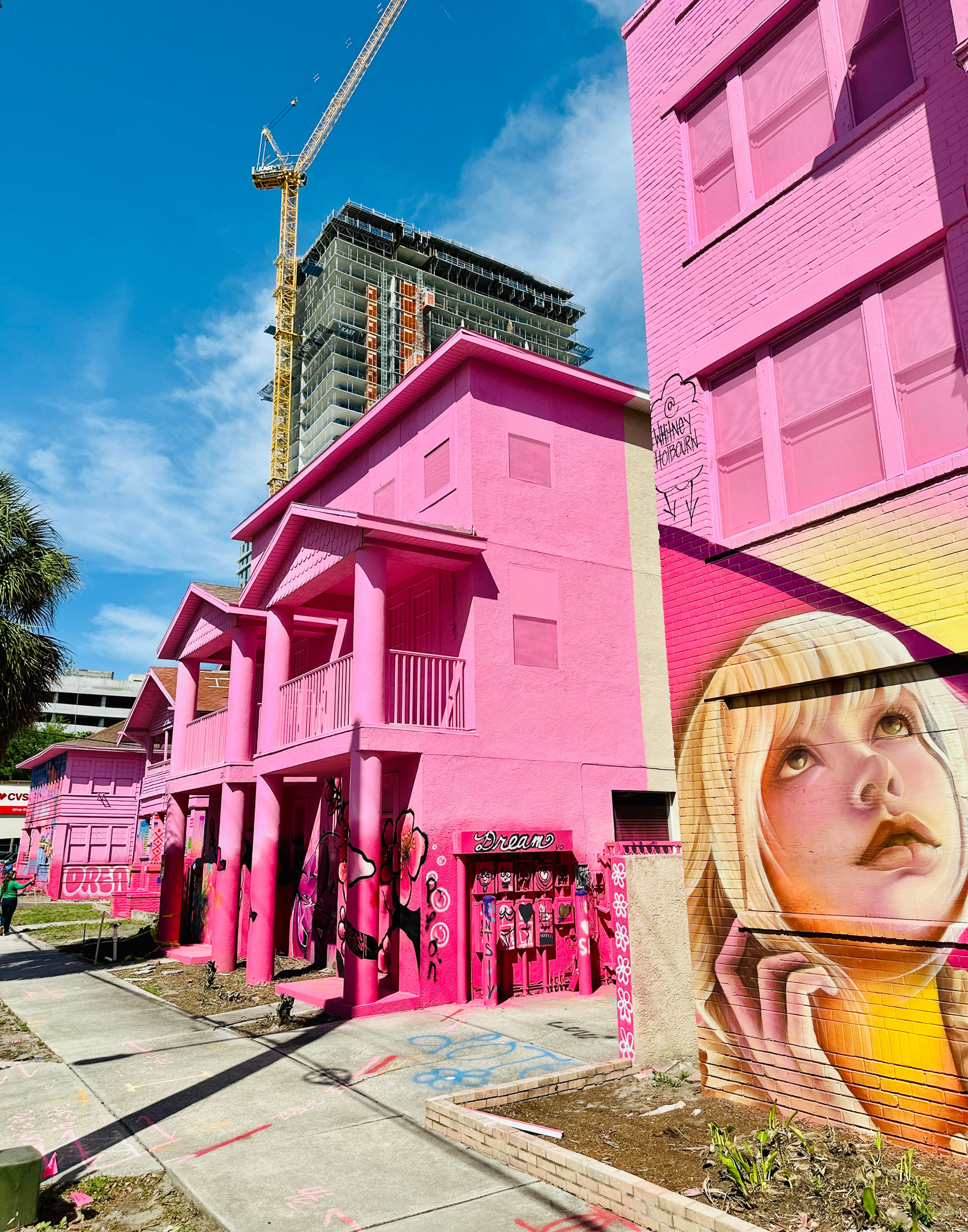 Bright pink buildings with murals, including a large portrait of a woman. A crane looms in the background. Clear blue sky overhead.