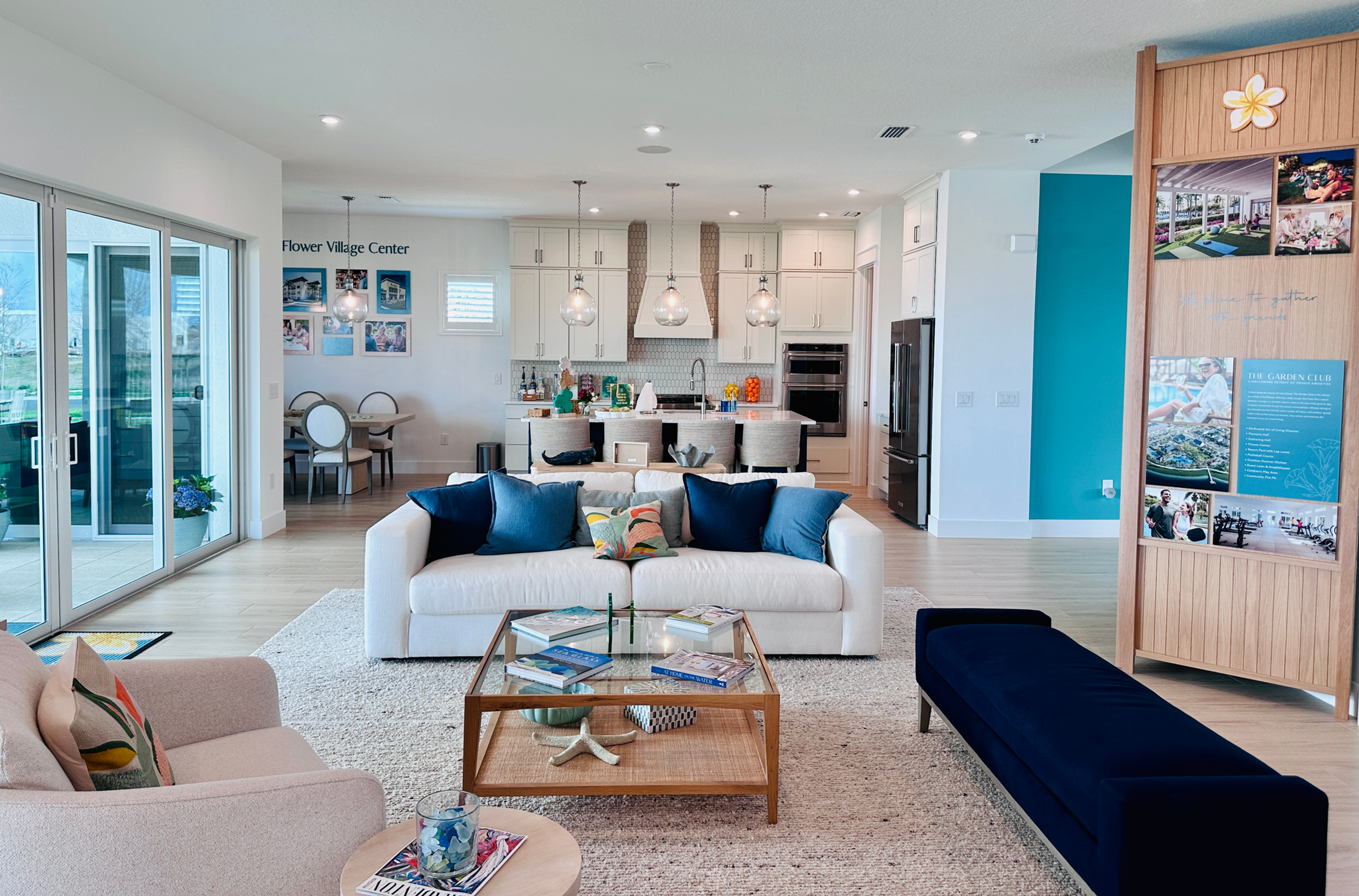Modern living room with white sofas, blue cushions, and a glass coffee table. Kitchen and dining area visible in background. Bright, airy space.