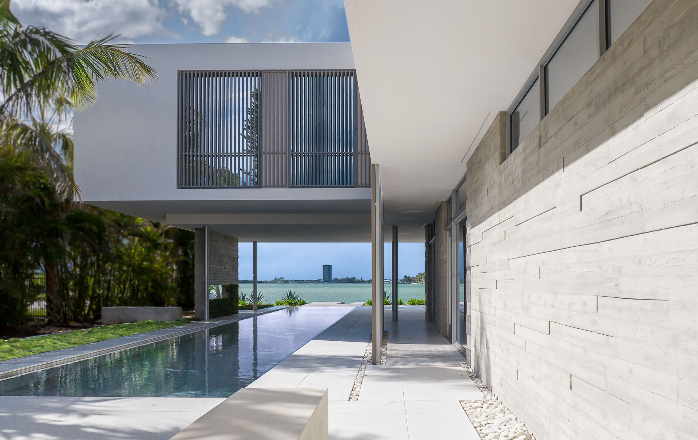 Modern waterfront home with reflective pool, white stone walls, palm trees, and ocean view under a bright sky.