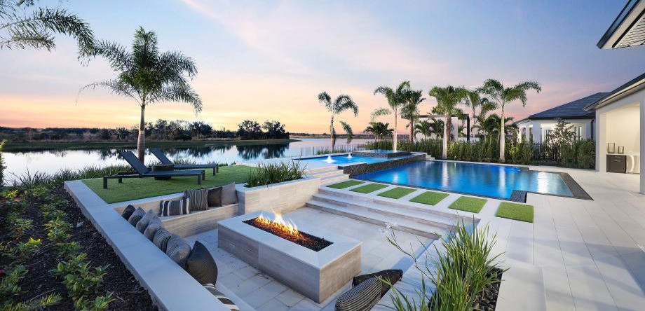Luxury backyard with blue pool, fire pit, lounge chairs and palm trees beside a calm lake at sunset.