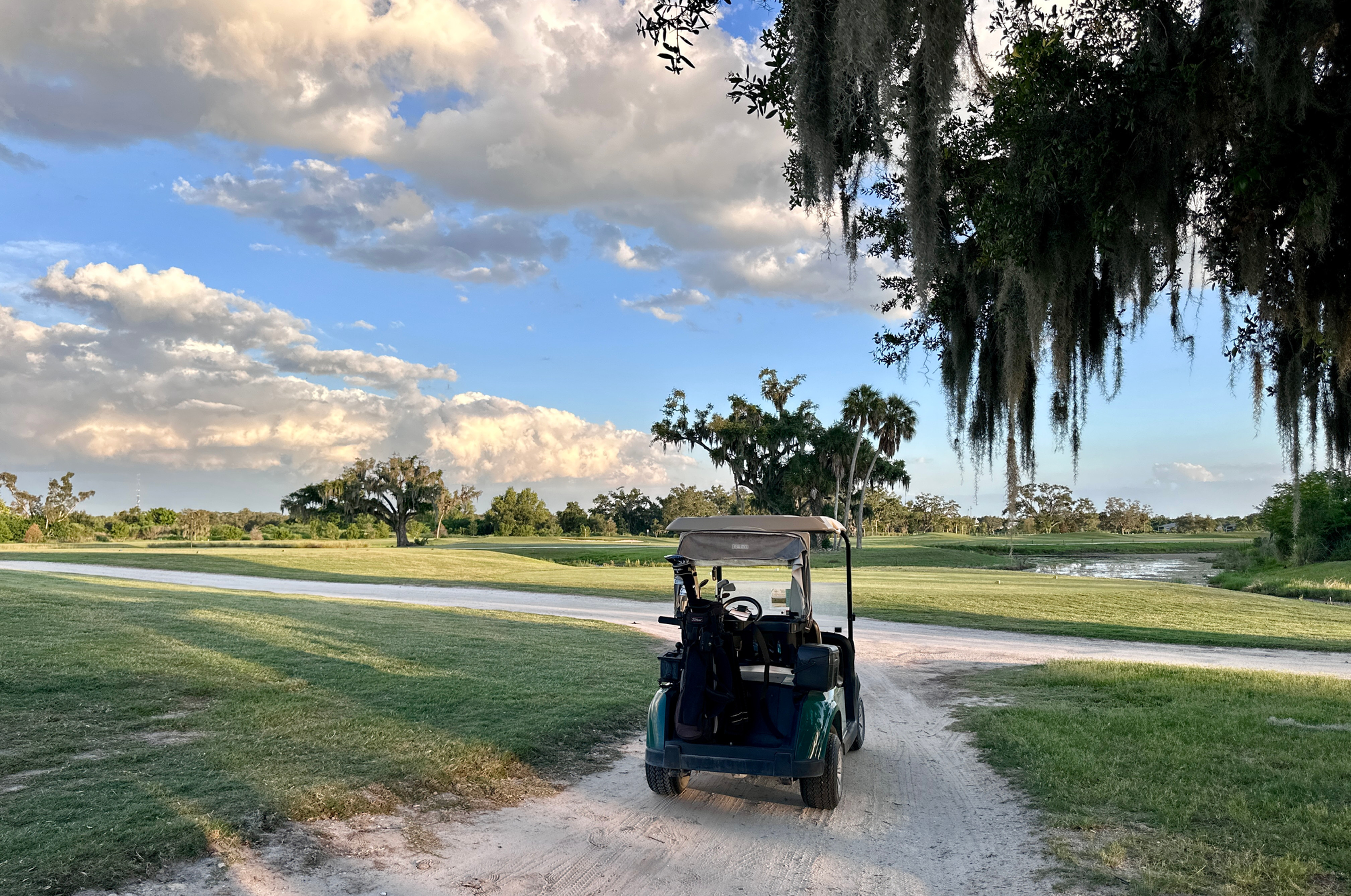 Golf cart on a sandy path at a quiet golf course, with grassy fairways, Spanish moss, and a cloudy blue sky.
