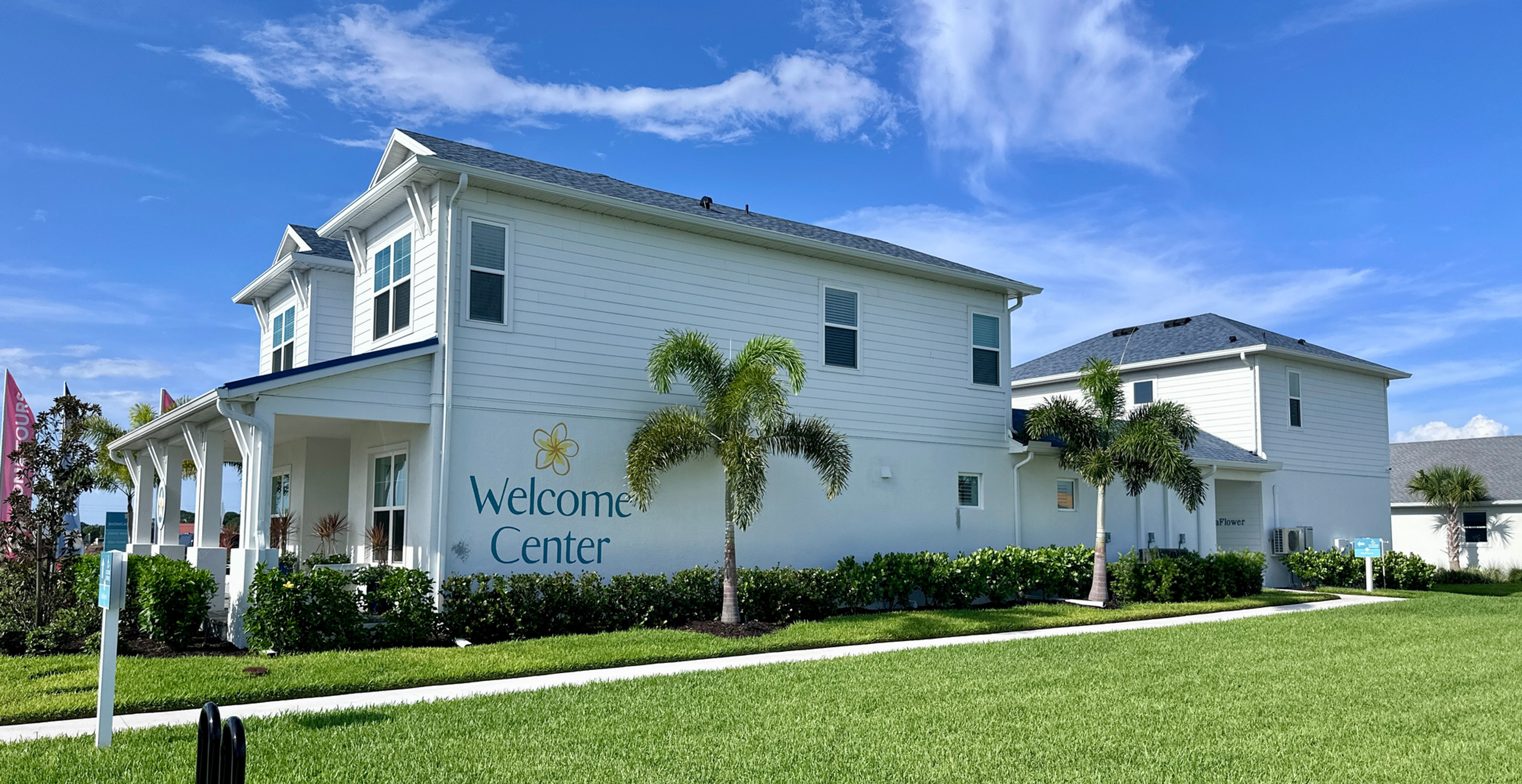 White Welcome Center building with palm trees and manicured lawn under a bright blue sky with wispy clouds. This is the Eventide model by Cardel Homes.